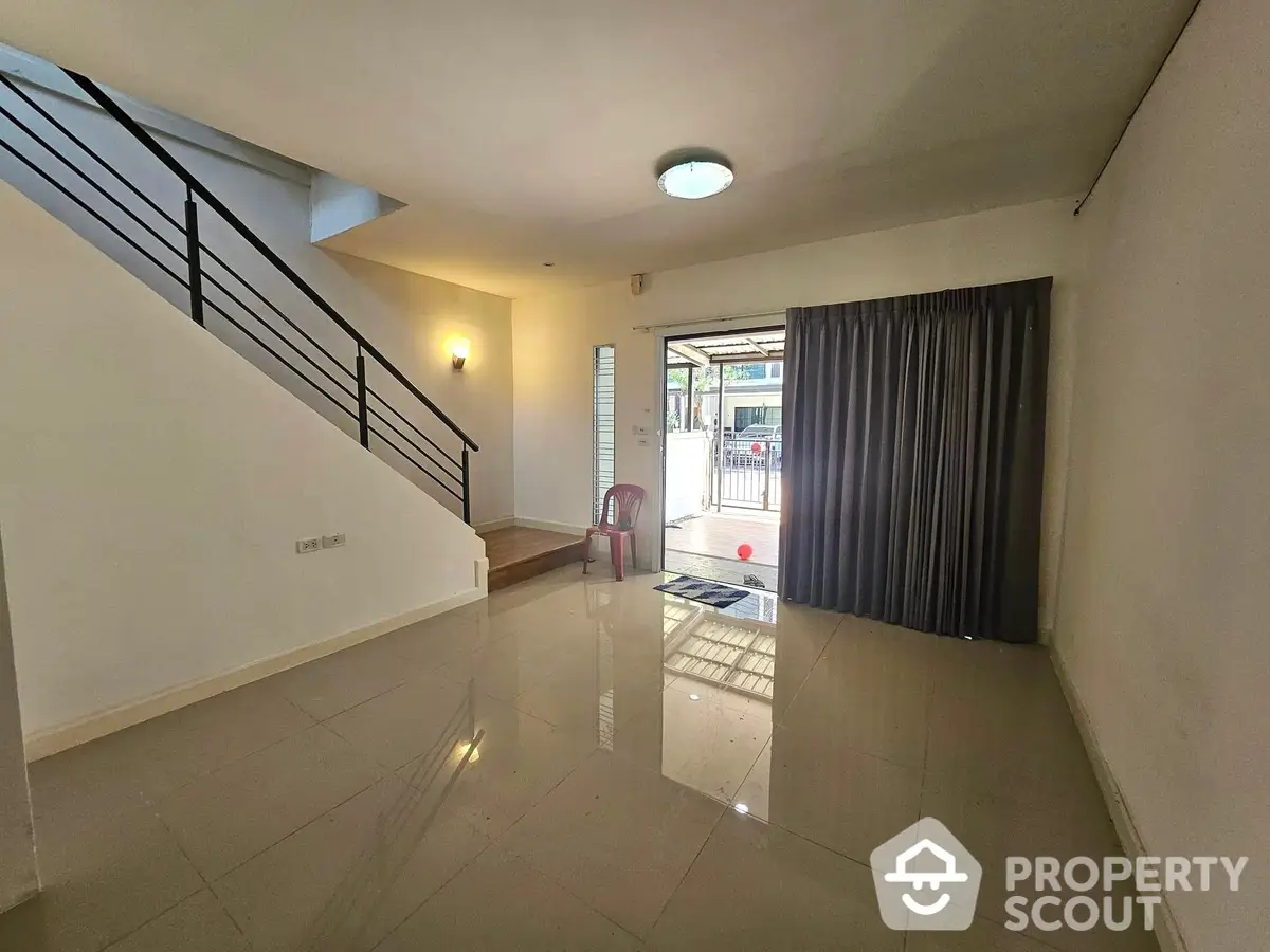 Spacious living room with glossy tiled flooring, elegant staircase, and ample natural light streaming through large glass doors leading to a cozy balcony.