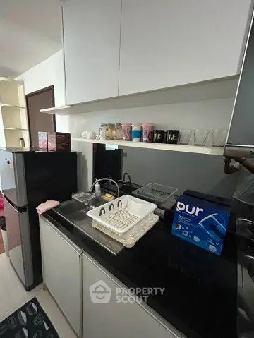 Modern kitchen with sleek black countertop and white cabinets, featuring a compact fridge and dish rack.