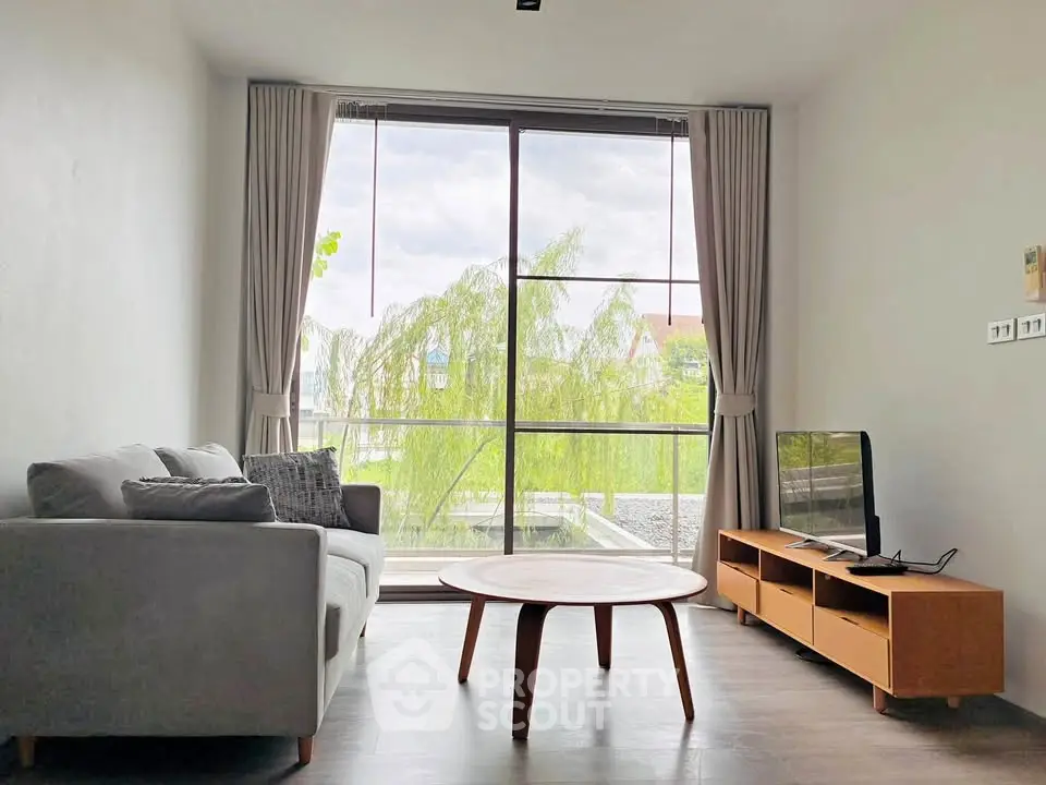 Modern living room with large window and stylish furniture, offering a serene view of greenery.