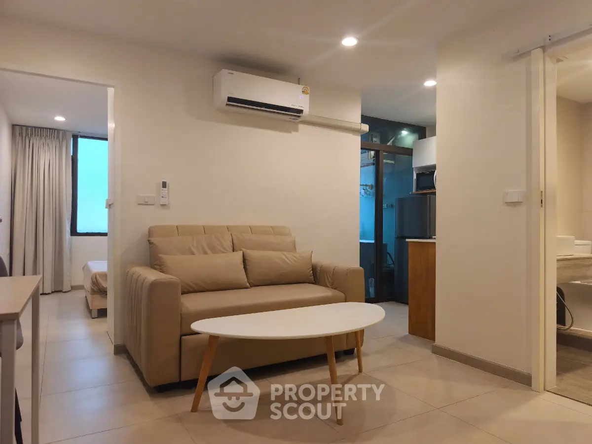 Modern living room with beige sofa, air conditioning, and open view to kitchen and bedroom.