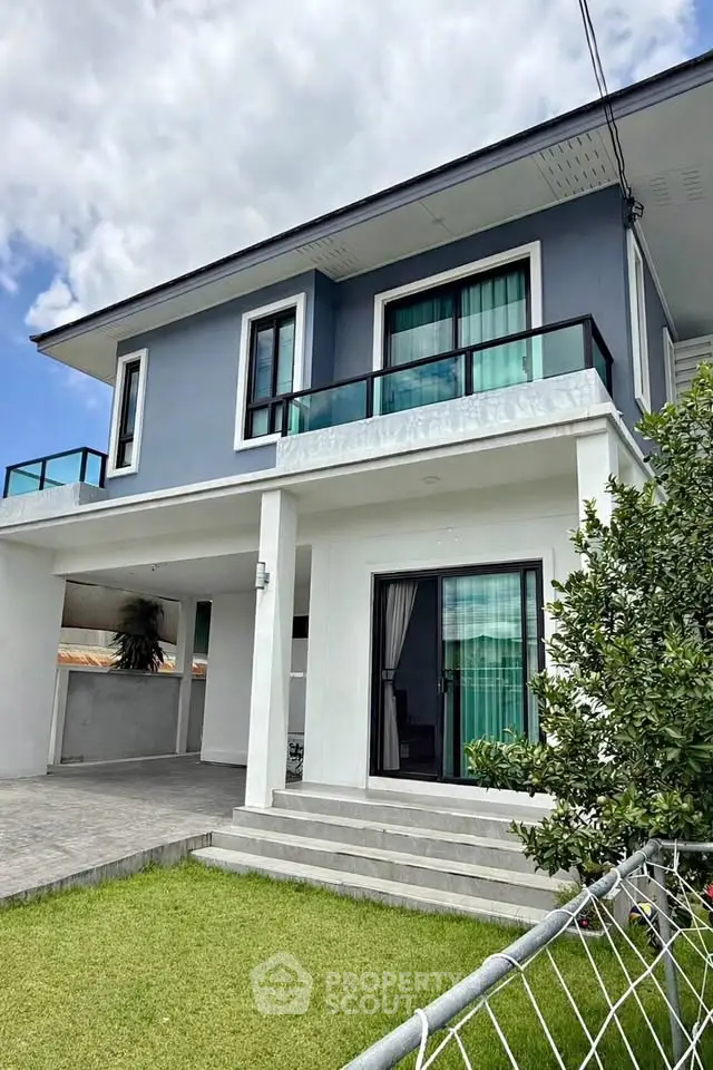 Modern two-story house with large windows and balcony, featuring a lush green lawn and sleek exterior design.