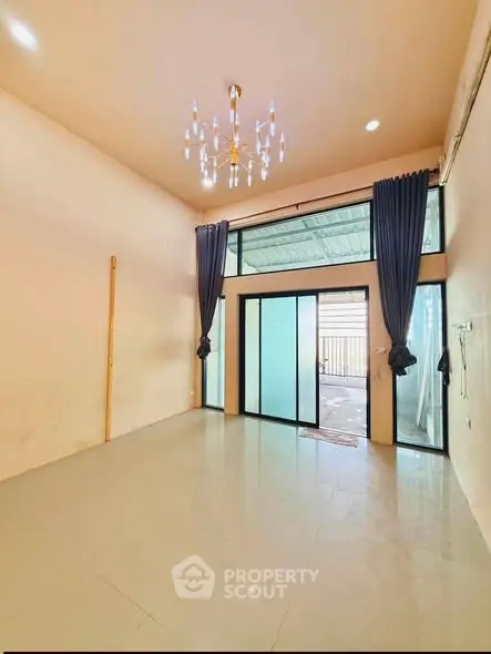Spacious living room with high ceiling and elegant chandelier, featuring large sliding glass doors leading to a balcony.
