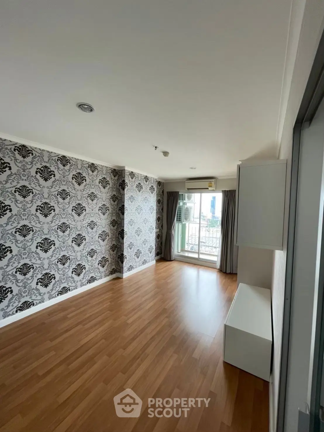 Spacious empty room with elegant wallpaper and wooden flooring, featuring a balcony view.