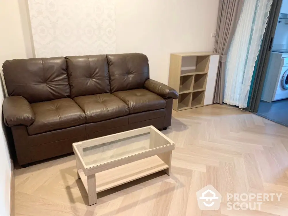 Cozy living room with brown leather sofa and glass coffee table, adjacent to a modern kitchen with washing machine.