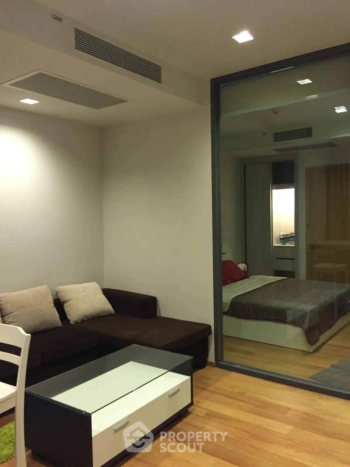 Modern living room with glass partition to bedroom, featuring sleek furniture and wooden flooring.