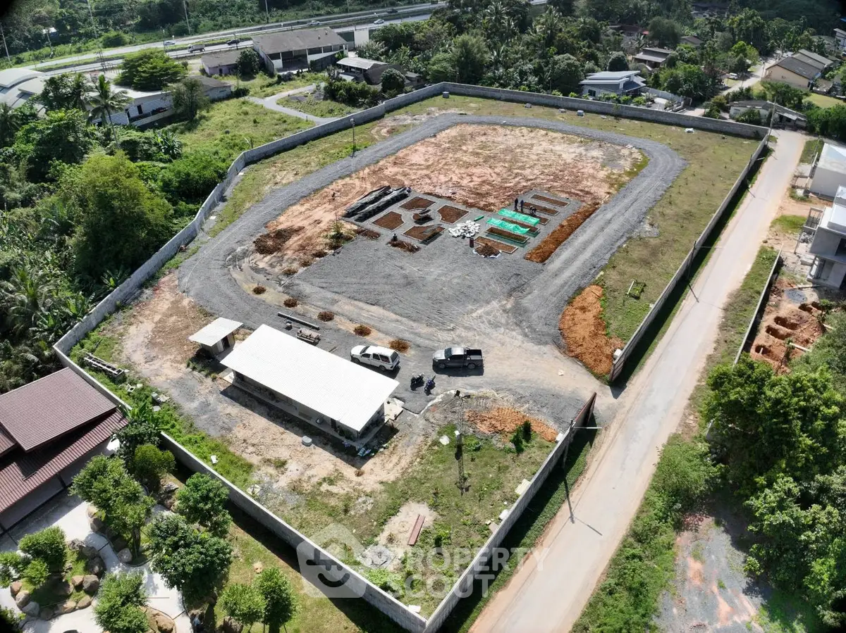 Aerial view of a large fenced construction site with surrounding greenery and nearby road access.