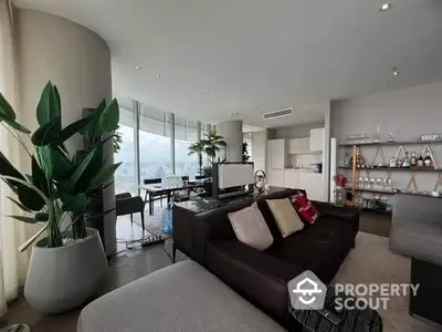 Spacious modern living room with large windows offering a panoramic city view, sleek furnishings, and an open-plan layout leading to a well-equipped kitchen.