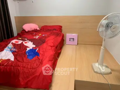 Cozy bedroom with vibrant red bedding and wooden flooring, featuring a standing fan for comfort.