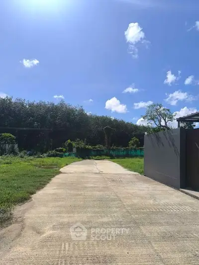Spacious driveway leading to a modern gate with lush greenery and clear blue sky.