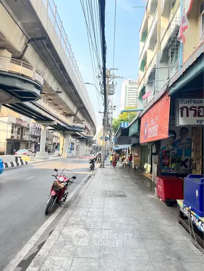 Bustling urban street with shops and elevated train tracks, vibrant city life.