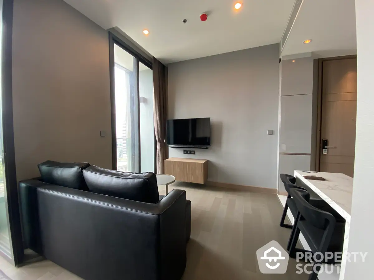 Modern living room with sleek black leather sofa and a flat-screen TV, complemented by natural light from floor-to-ceiling windows, offering a cozy urban retreat.