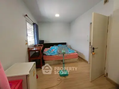Cozy bedroom with wooden flooring, bed, fan, and natural light from window.