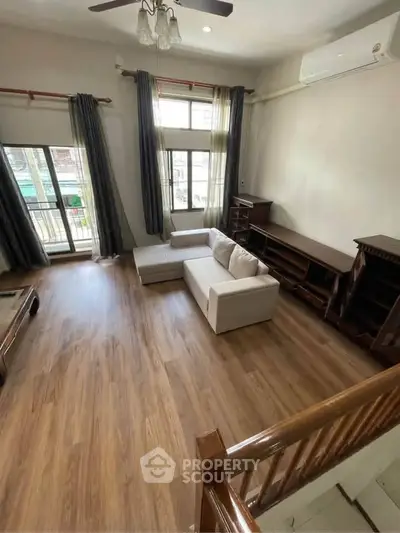 Spacious living room with wooden flooring and modern furniture, featuring large windows for natural light.