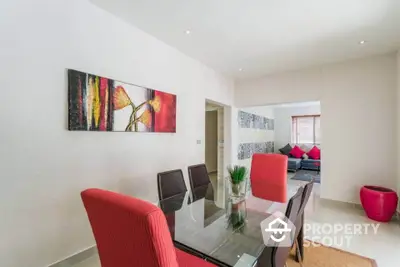 Modern dining area with glass table and red chairs, featuring contemporary artwork and open layout to living room.