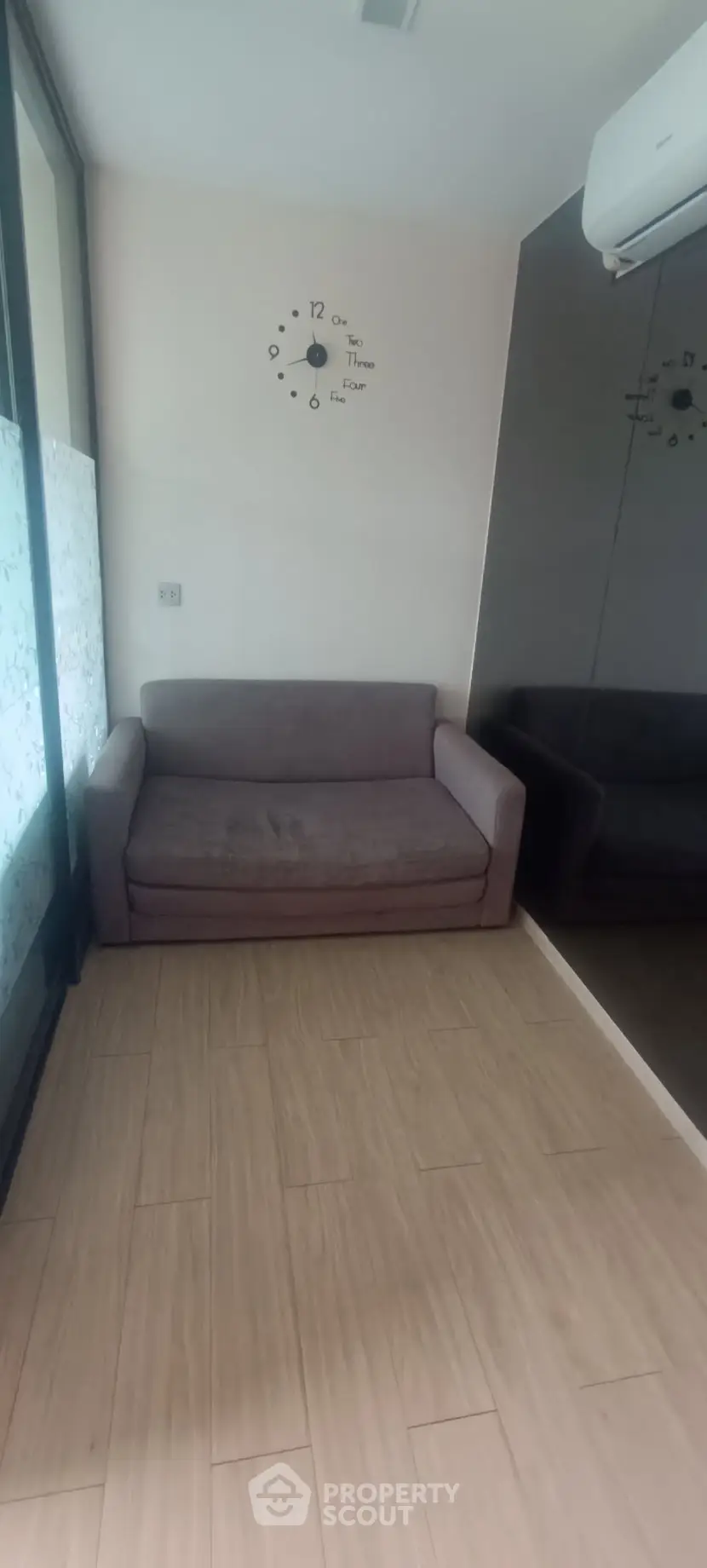 Cozy small living room with a sofa and wall clock, featuring wooden flooring and mirrored wall for spacious feel.