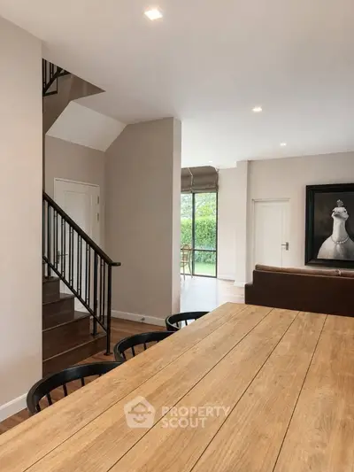 Spacious modern living room with wooden dining table and staircase, featuring large windows and elegant decor.