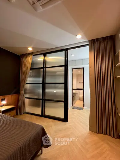 Modern bedroom with sliding glass door leading to a stylish bathroom, featuring elegant lighting and wooden flooring.