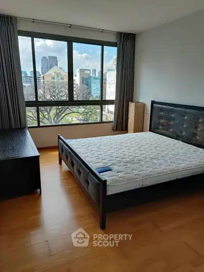 Spacious bedroom with large window offering stunning city views, featuring a stylish bed and wooden flooring.