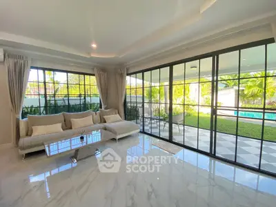 Spacious living room with large windows overlooking a lush garden and pool, featuring elegant marble flooring.