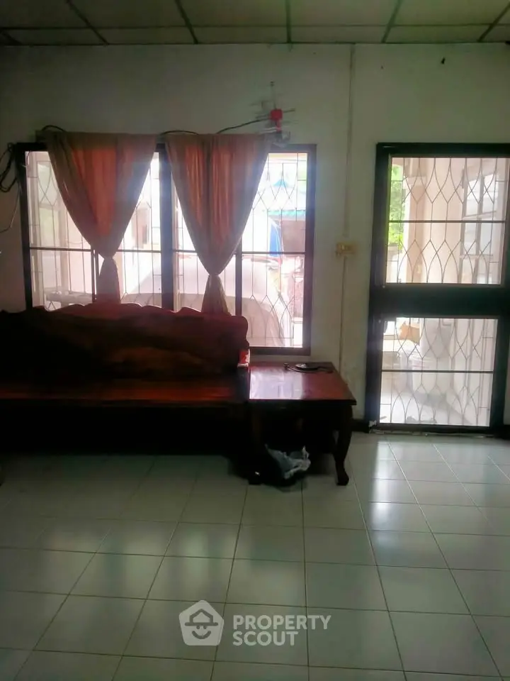 Cozy living room with natural light and tiled flooring, featuring a wooden sofa and large windows.
