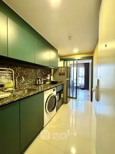 Modern kitchen with sleek green cabinets, washing machine, and open layout leading to a bright living area.