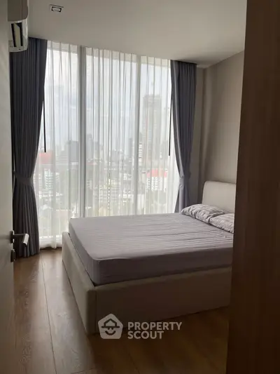 Modern bedroom with floor-to-ceiling windows and city view, featuring elegant curtains and wooden flooring.