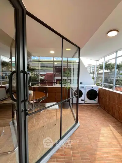 Modern balcony with glass doors, seating area, and laundry appliances in urban setting.