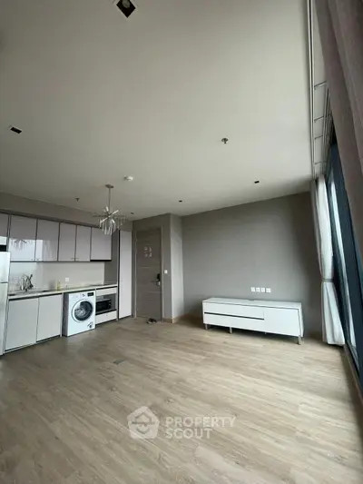 Modern open-plan living area with kitchen and washing machine, featuring large windows and wooden flooring.