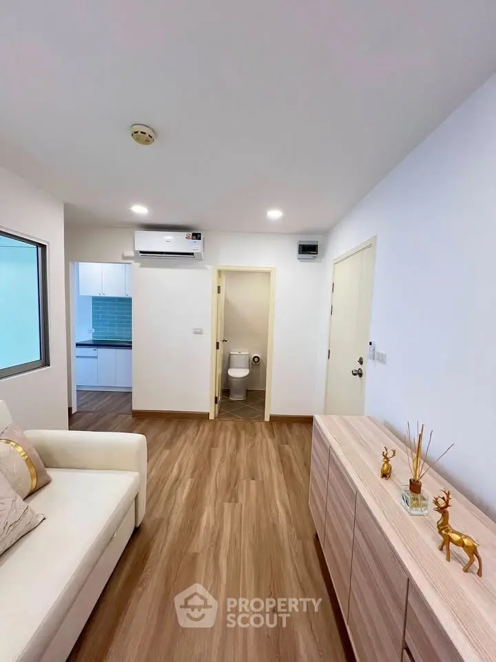 Modern living room with wooden flooring, white sofa, and sleek cabinetry leading to a kitchen and bathroom.