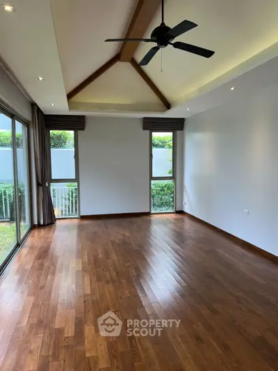Spacious living room with wooden flooring and ceiling fan, large windows overlooking greenery.