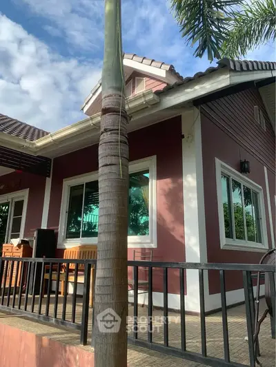 Charming single-story house with red exterior and white trim, surrounded by lush greenery and palm trees.