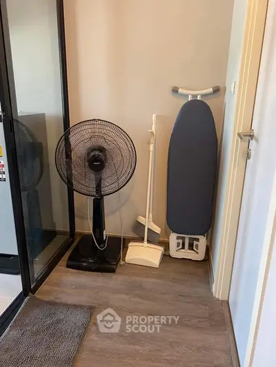 Compact storage corner with fan, ironing board, and cleaning tools on wooden floor.