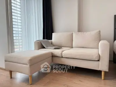 Modern living room with beige sofa and ottoman, featuring floor-to-ceiling windows and wooden flooring.