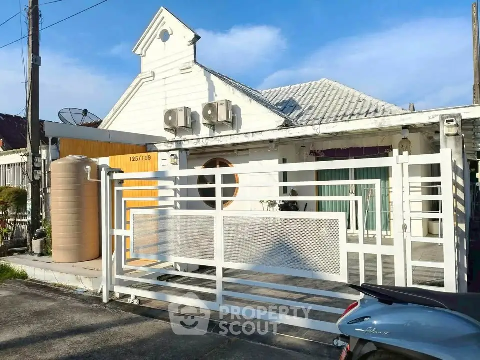 Charming white house with unique architecture and gated entrance, featuring air conditioning units and a water tank.