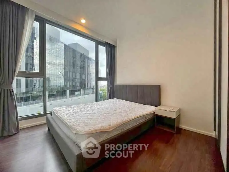 Modern bedroom with large window and city view, featuring sleek furniture and hardwood floors.