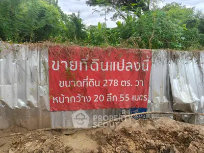 Vacant land for sale with a red banner displaying plot size and dimensions, surrounded by lush greenery.