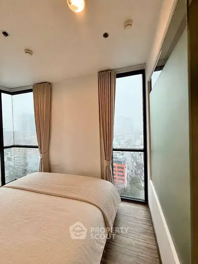 Modern bedroom with large windows offering city views, featuring elegant curtains and cozy bedding.