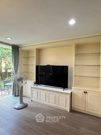 Spacious living room with built-in shelves and large TV, overlooking a lush garden view.
