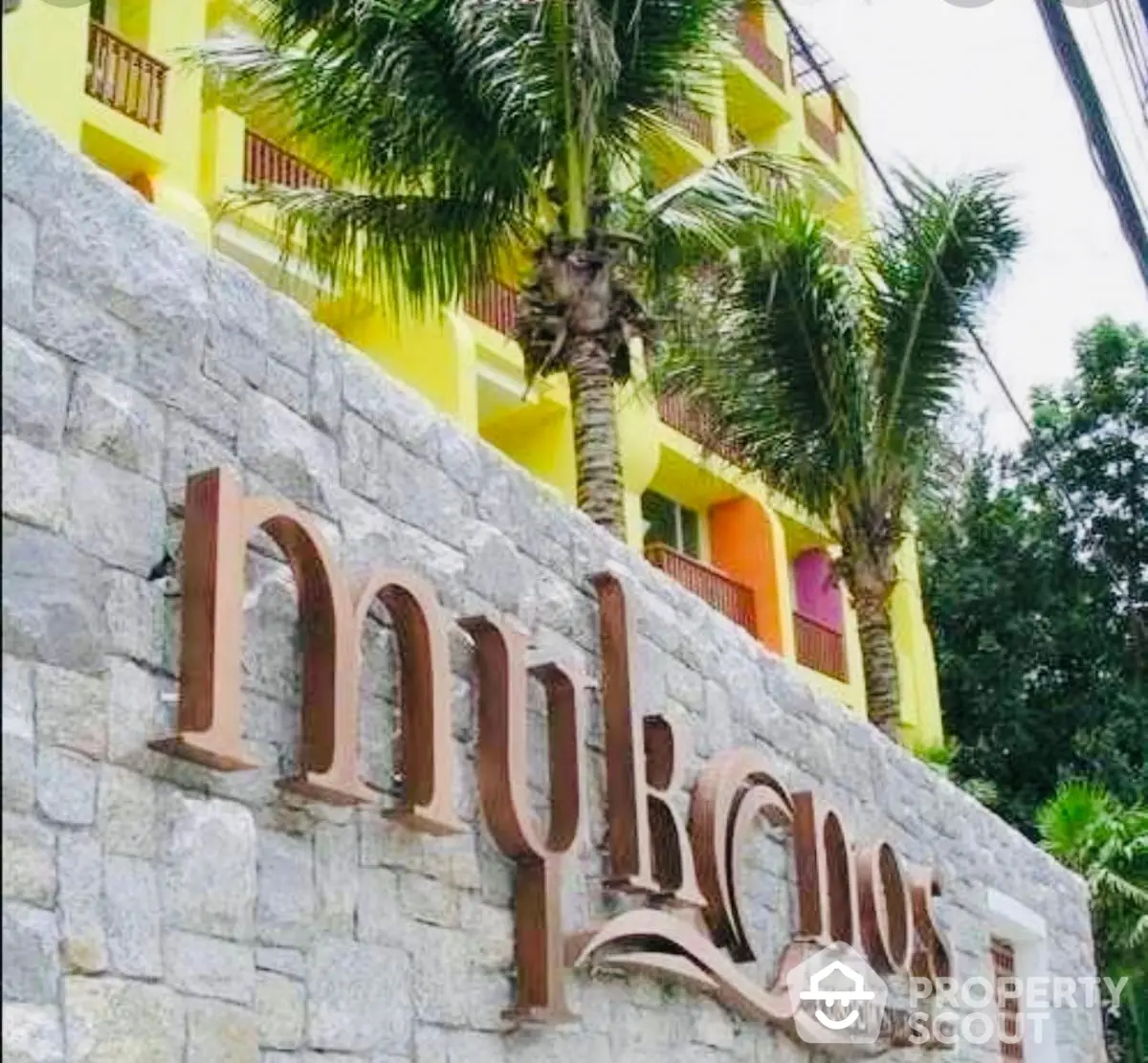 Vibrant Mykonos condominium exterior with palm trees and stone wall signage.