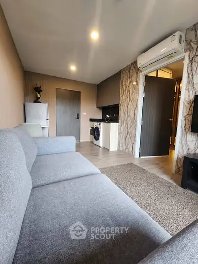 Modern living room with gray sofa, air conditioning, and compact kitchen featuring washing machine.