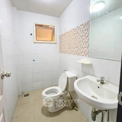 Compact bathroom with white tiles and modern fixtures, featuring a toilet, sink, and small window for natural light.