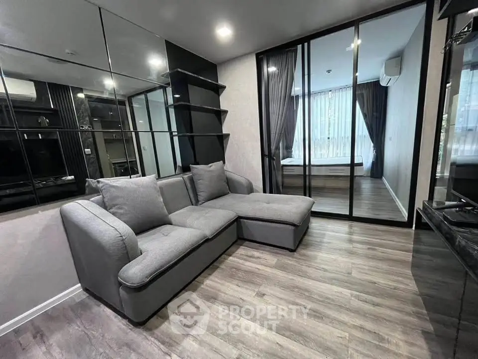 Modern living room with sleek gray sofa and mirrored wall, leading to a stylish bedroom with floor-to-ceiling windows.