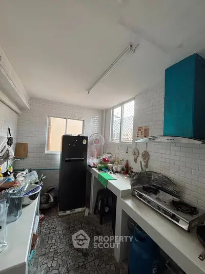 Compact kitchen with gas stove and fridge, featuring white tiled walls and natural light from window.