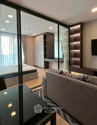 Modern open-plan living room with glass partition to bedroom, featuring sleek furniture and ample natural light.
