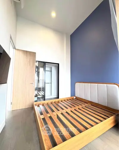 Modern bedroom with high ceiling and wooden bed frame, featuring a blue accent wall and natural light.