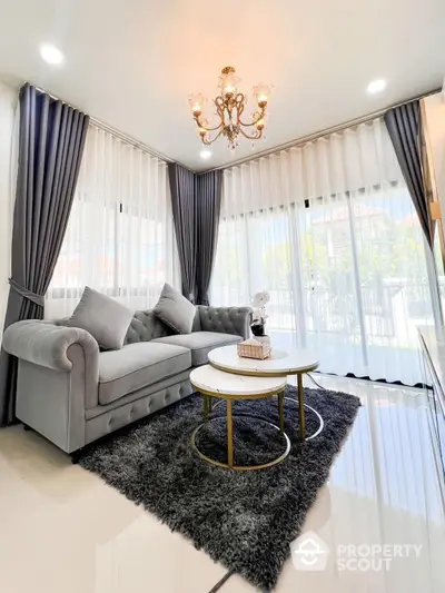 Elegant living room with plush gray sofa, chic marble-top coffee table, luxurious chandelier, and floor-to-ceiling windows draped in stylish curtains, offering a cozy yet sophisticated space.