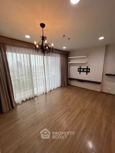 Spacious living room with elegant chandelier, large windows draped in sheer curtains, and warm wooden flooring, perfect for modern living.