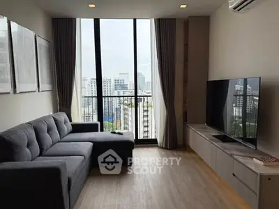 Modern living room with large windows and city view, featuring a stylish gray sofa and flat-screen TV.