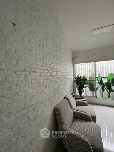 Modern living room with white brick wall and cozy seating area, featuring large windows and lush greenery.