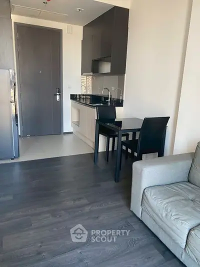 Modern apartment interior with compact kitchen and cozy living area, featuring sleek cabinetry and dark wood flooring.